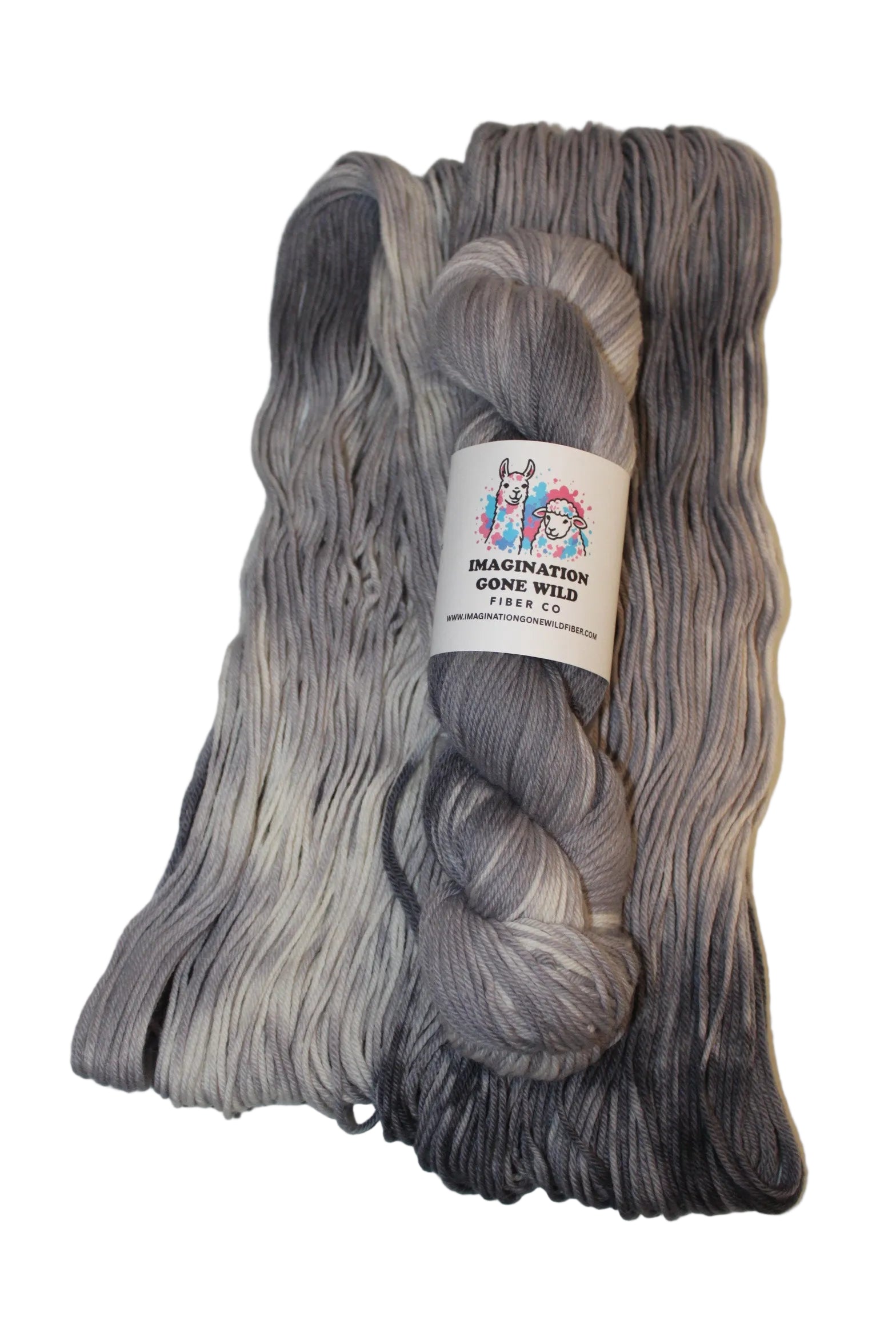 A skein of variegated yarn with a white paper label, placed on top of a folded matching yarn bundle. The yarn has a color-shifting gradient from light silver-gray to deep charcoal black.